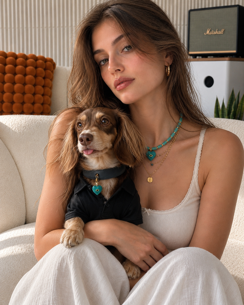 A stylish woman and her dog sitting on a white sofa, both wearing matching Muttu turquoise crystal necklaces. The dog is also wearing a black pet shirt, showcasing the luxury The Matching Bond collection.